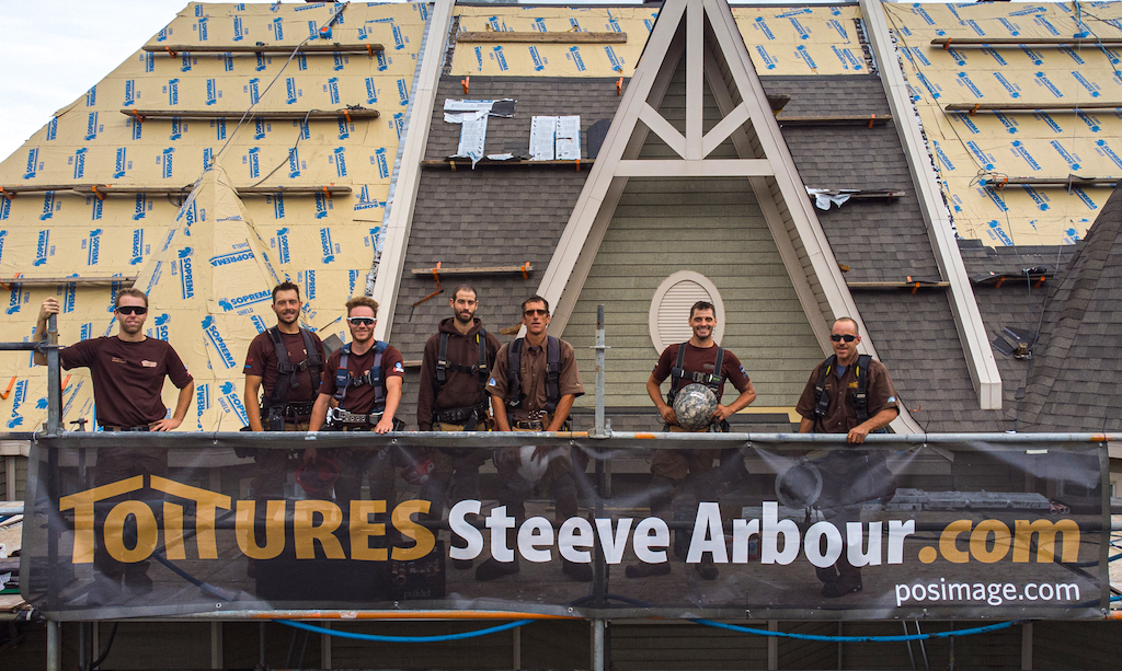 Équipe de Toitures Steeve Arbour sur le toît en construction d'un immeuble, avec grosse banderole affichant le nom de l'entreprise accrochée sur le toît.